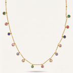 A gold chain necklace featuring multiple small round bezel-set gemstones in green, pink, blue, and clear, displayed against a light cream background.