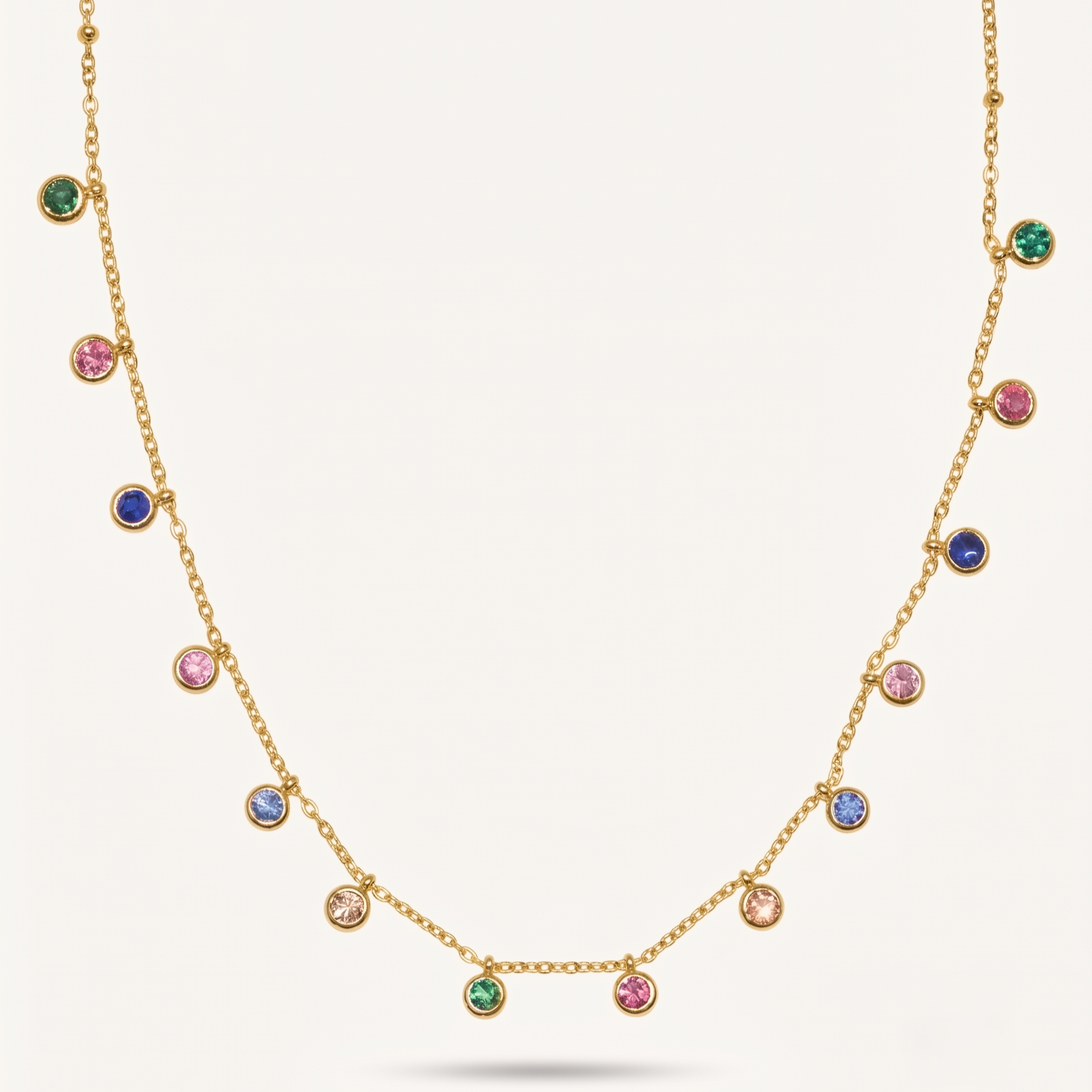 A gold chain necklace featuring multiple small round bezel-set gemstones in green, pink, blue, and clear, displayed against a light cream background.